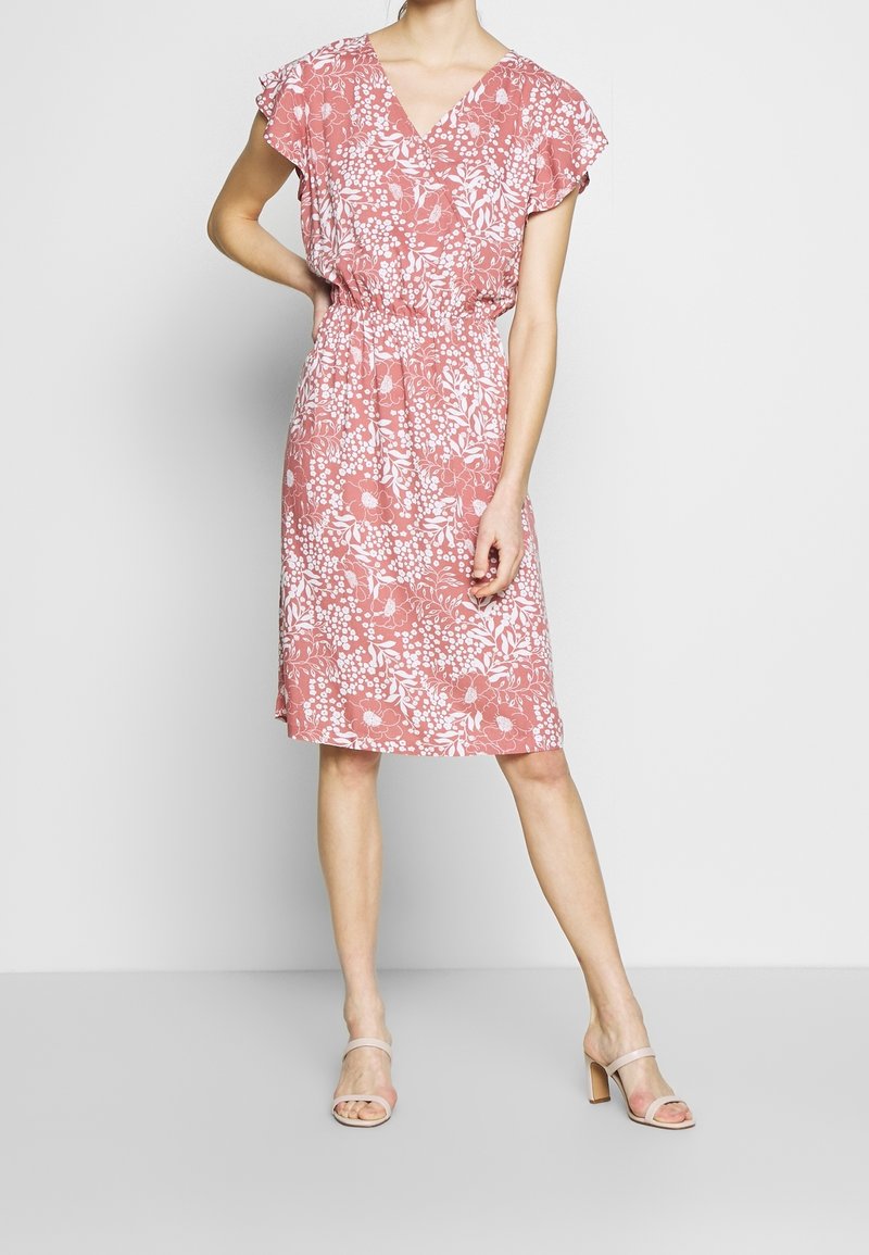 Femme portant une robe rose mi-longue avec imprimé floral blanc et manches courtes, associée à des sandales à talons beiges à bouts ouverts.