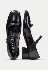 Zapatos Mary Jane de charol negro con puntera redondeada, correa con hebilla y tacón texturizado. Presentan un logo en relieve en la plantilla.