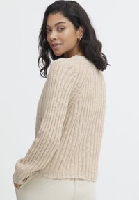Pull en maille beige avec une texture côtelée, coupe décontractée, manches longues et une taille légèrement cropped. Il possède un col rond et est fabriqué dans un matériau doux.