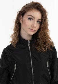 Giacca nera con zip e zip metallici, colletto alto e texture lucida. Il modello presenta capelli mossi e trucco delicato.