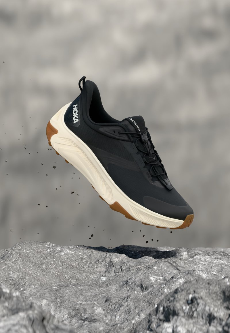 Crno-bijela Hoka tenisica za trčanje lebdi u zraku iznad kamenitog tla s malim komadićima koji lete oko nje.