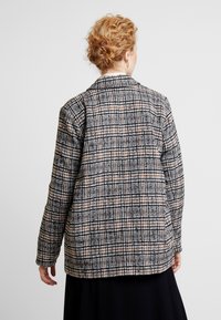 Kostkovaný blazer v černobílé a hnědé barvě. Disponuje strukturovaným střihem, klopovým límcem a dlouhými rukávy s hladkou texturou.