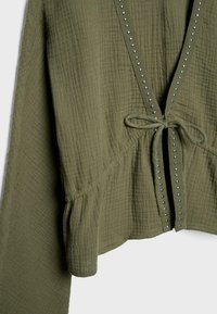 Veste en tissu texturé vert olive avec manches longues, bordure cloutée le long de l'ouverture avant, et un nœud noué à la taille.