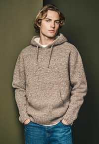 Brun strikket hættetrøje med en struktureret finish, der har en frontlomme, justerbar snor og ribbet manchetter. Båret over en hvid skjorte og jeans.