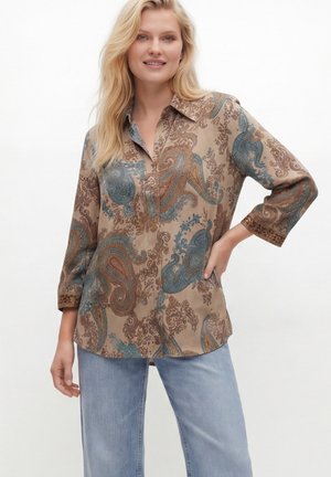 Blonde woman wearing a brown paisley-patterned long-sleeve shirt and light blue jeans, standing with one hand on hip.