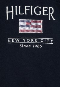 Tessuto di cotone navy con testo ricamato bianco "HILFIGER", un design a bandiera e "NEW YORK CITY Dal 1985" in filo bianco.