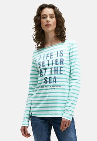 Frau mit lockigem Haar, die ein langärmliges, weiß-tee-farbenes gestreiftes Shirt mit dem Text "Das Leben ist besser am Meer" und blaue Jeans trägt.