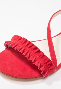 Sandale ouverte en suède rouge avec une bride à volants sur les orteils et une fine bride à la cheville sur fond blanc.
