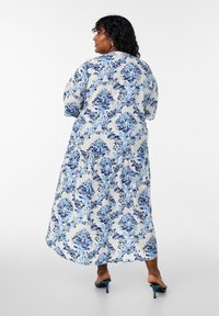 Robe maxi légère à imprimé floral en blanc et bleu. Présente des manches bouffantes, un ourlet évasé et une texture douce. Design à dos fermé.
