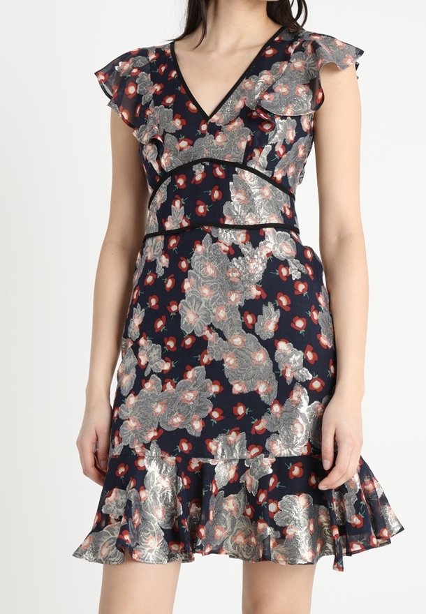 Femme portant une robe bleu marine avec des motifs floraux argentés et rouges, des manches flottantes, un col en V et un ourlet volanté.