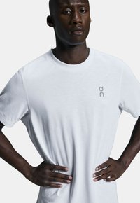 Weißes Baumwoll-T-Shirt mit rundem Ausschnitt und kurzen Ärmeln, mit einem kleinen grauen Logo auf der linken Brust. Glatte Textur, lockere Passform.