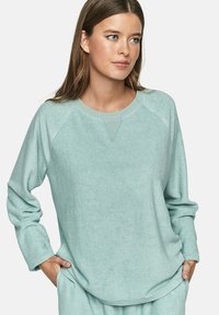 Femme aux longs cheveux bruns portant un haut à manches longues doux de couleur bleu clair et un bas assorti, les mains dans les poches, regardant à droite.