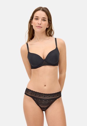 Mujer con un sujetador acolchado negro con detalles de encaje y bragas a juego de encaje negro, de pie contra un fondo claro y sencillo.