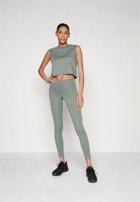 Kvinne står med den ene hånden på hoften iført ermeløs crop top, høytlivte leggings og svarte joggesko mot en ensfarget bakgrunn.