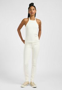 Vit, ribbad halterneck-topp i kombination med åtsittande vita jeans och beige sneakers. Modellen står med avslappnade armar och visar upp outfitens stilrena design.