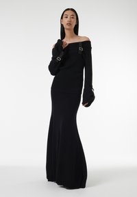 Zwarte gebreide trui met een off-shoulder ontwerp, gekenmerkt door lange mouwen met een gespaccent. Gepaarde met een lange, strakke zwarte rok.