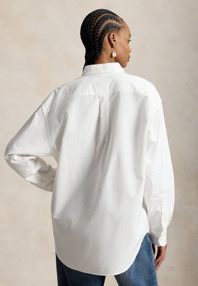 Frau mit Cornrow-Zöpfen steht mit dem Rücken zur Kamera, trägt ein locker sitzendes weißes Hemd mit Knöpfen und blaue Jeans vor einem einfarbigen Hintergrund.