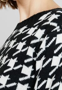 Svetr s černobílým houndstooth vzorem pletený s žebrovaným výstřihem, který má texturovaný vzor se střídajícími se tvary. Detailní pohled.