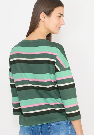 Femme aux longs cheveux bruns portant une chemise rayée verte avec des bandes roses, blanches et vert foncé, associée à un jean en denim bleu.