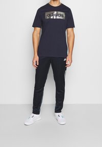 Camiseta azul marino con logo metálico plata de "adidas", combinada con pantalones de chándal oscuros con acentos de panel lateral. Zapatillas blancas completan el conjunto.