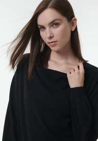 Zwarte trui met een off-the-shoulder ontwerp, zachte textuur en lange mouwen. Model past de stof bij de halslijn aan, voor een effen achtergrond.