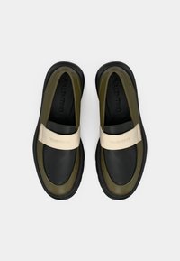 Zapatillas negras y verde oliva sin cordones con una tira beige que lleva el logo "Valentino", superficies lisas y un diseño de punta redonda.