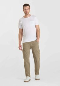Man som står och bär en vit t-shirt med bröstficka, beiga cargo-byxor och vita sneakers framför en plain ljus bakgrund.