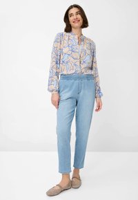 Blusa a fantasia floreale in pesca chiaro con bordi blu, abbinata a jeans tapered in azzurro chiaro con vita elastica e scarpe basse beige.