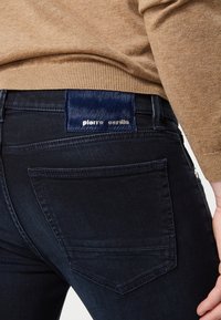 Černé džíny z jeanu mají přiléhavý střih, klasický pětikapsový styl a štítek s námořnicky modrými a kovovými akcenty na zadním pásu.