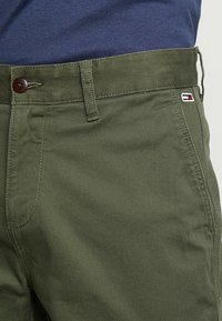 Oliivivihreät puuvillachinot, joissa on tekstuuri, ruskea nappikiinnitys ja pieni logo-uutus punaisella, valkoisella ja sinisellä.