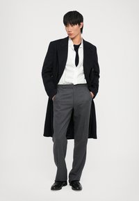 Lungo cappotto blu navy sopra una camicia bianca e una cravatta nera; pantaloni grigi su misura; scarpe nere; design elegante e moderno, con texture morbide.