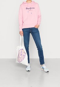 Persona vestida con una sudadera rosa, jeans azules y zapatillas, sosteniendo una bolsa de tote blanca con un diseño púrpura de "Paz Amor".