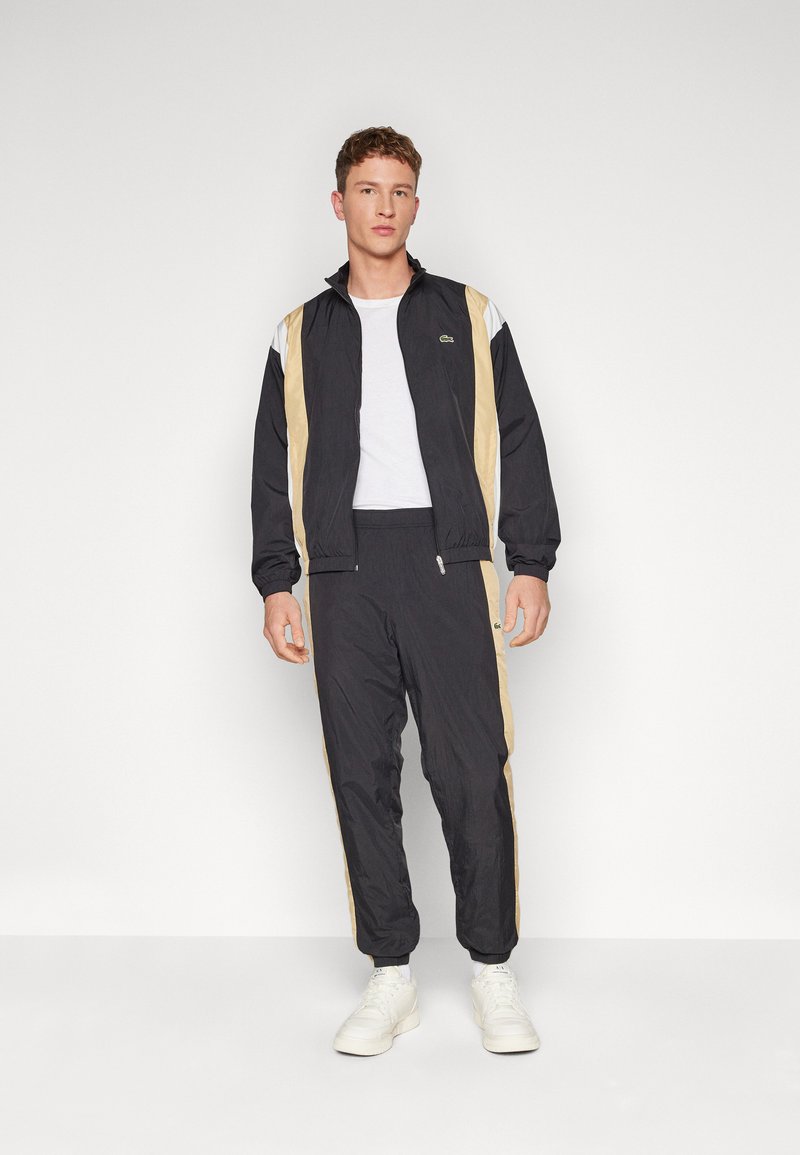 Survêtement noir avec des rayures latérales crème et blanches, veste à zip, pantalon avec taille élastique et un petit logo sur la veste.