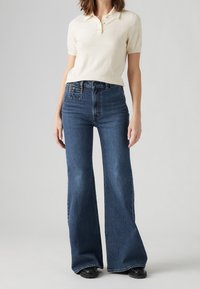 Ljus beige kortärmad polo skjorta kombinerad med högmidjade, utsvängda mörkblå jeans med en strukturerad yta och knappstängning i midjan.