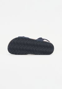 Marinblå sandal med en texturerad gummisula som har ett vågigt mönster. Justerbara remmar ger en säker passform. Sulan erbjuder grepp och hållbarhet.