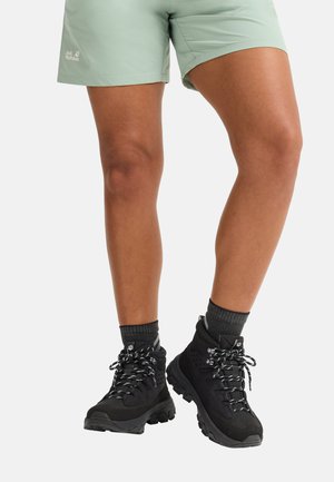 Jambes portant un short vert clair, des chaussettes gris foncé et des chaussures de randonnée noires avec des lacets à motifs sur fond blanc.