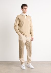 Polo bej cu mânecă lungă, cu guler alb și logo, purtat cu pantaloni bej cu accente de catifea și buzunare laterale, completat cu adidași albi.