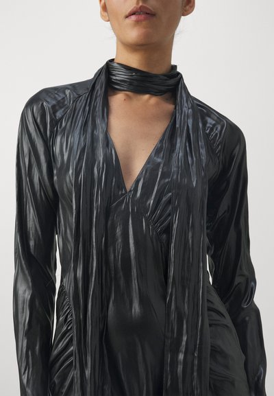 Robe noire, brillante, à manches longues avec un décolleté en V profond et un détail de foulard torsadé, présentant une texture drapée et des côtés froncés.