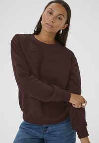Sweatshirt bordeaux avec le "27" en relief sur le devant, col rond, bords côtes au niveau des poignets et de l'ourlet, associé à un jean bleu. Texture du tissu douce.