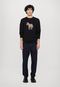 Black sweatshirt with a colorful horse graphic, ribbed cuffs, and hem. Paired with navy pants and black shoes, standing against a neutral backdrop.
