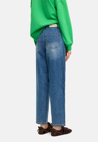 Niebieskie dżinsy z wysokim stanem, w odcieniu z wyblakłym efektem, o prostym kroju, z widocznymi tylnymi kieszeniami, zestawione z zielonym swetrem i brązowym obuwiem.