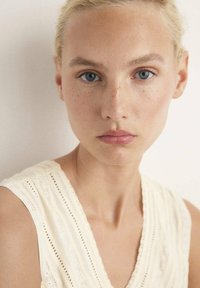 Primer plano de una persona de piel clara con pecas, que lleva una blusa texturizada color crema con rayas verticales y un escote en V.