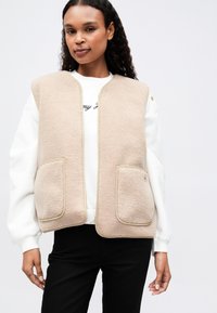 Vest din fleece bej cu o suprafață texturată, având două buzunare frontale, margini piped și un fermoar frontal. Purtați peste un hanorac alb.