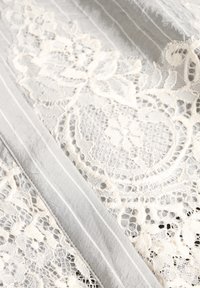 Tela de encaje en color crema con intrincados patrones florales, superpuesta sobre un fondo texturizado gris con finas rayas verticales.