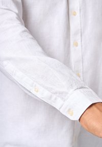 Camicia bianca di lino con maniche lunghe, bottoni in materiale chiaro, caratterizzata da una sottile texture e dettaglio ricamato sul polso.