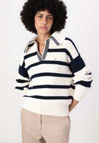 Pulover striped cu culorile navy și crem, guler în V, manșete și tiv ribbat, având un mic logo pe piept. Textură moale de tricot.