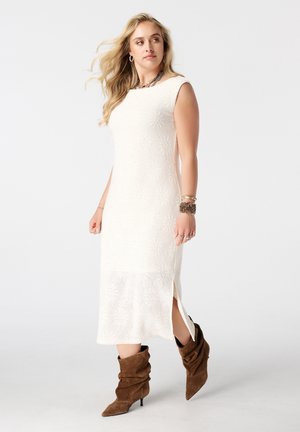 Vrouw met lang blond haar die een mouwloze witte midi-jurk met een zijsplit en bruine slappe laarzen met hakken draagt, kijkt opzij.