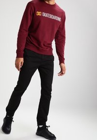 Burgunderfärgad sweatshirt med "SKATEBOARDING" grafik i gult, kombinerad med svarta byxor. Modellen har på sig svarta sportskor. Slät textur.