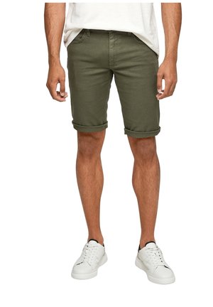 Mand iført op rullede olivengrønne shorts, hvide sneakers og en hvid t-shirt, stående med hænder afslappet ned langs siderne.