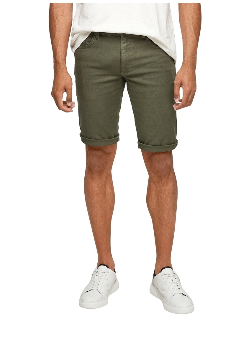 Homme portant un short olive retroussé, des baskets blanches et un t-shirt blanc, debout les mains détendues le long du corps.